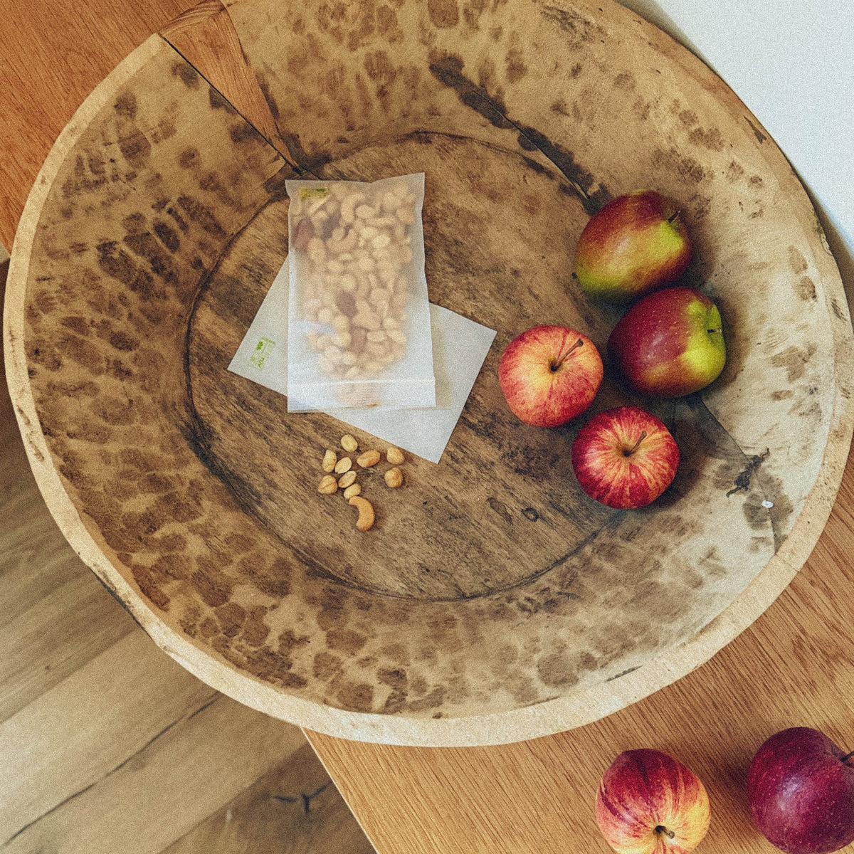 Top view of a rustic wooden bowl filled with a transparent bag of mixed nuts, a few individual nuts, and three red apples.